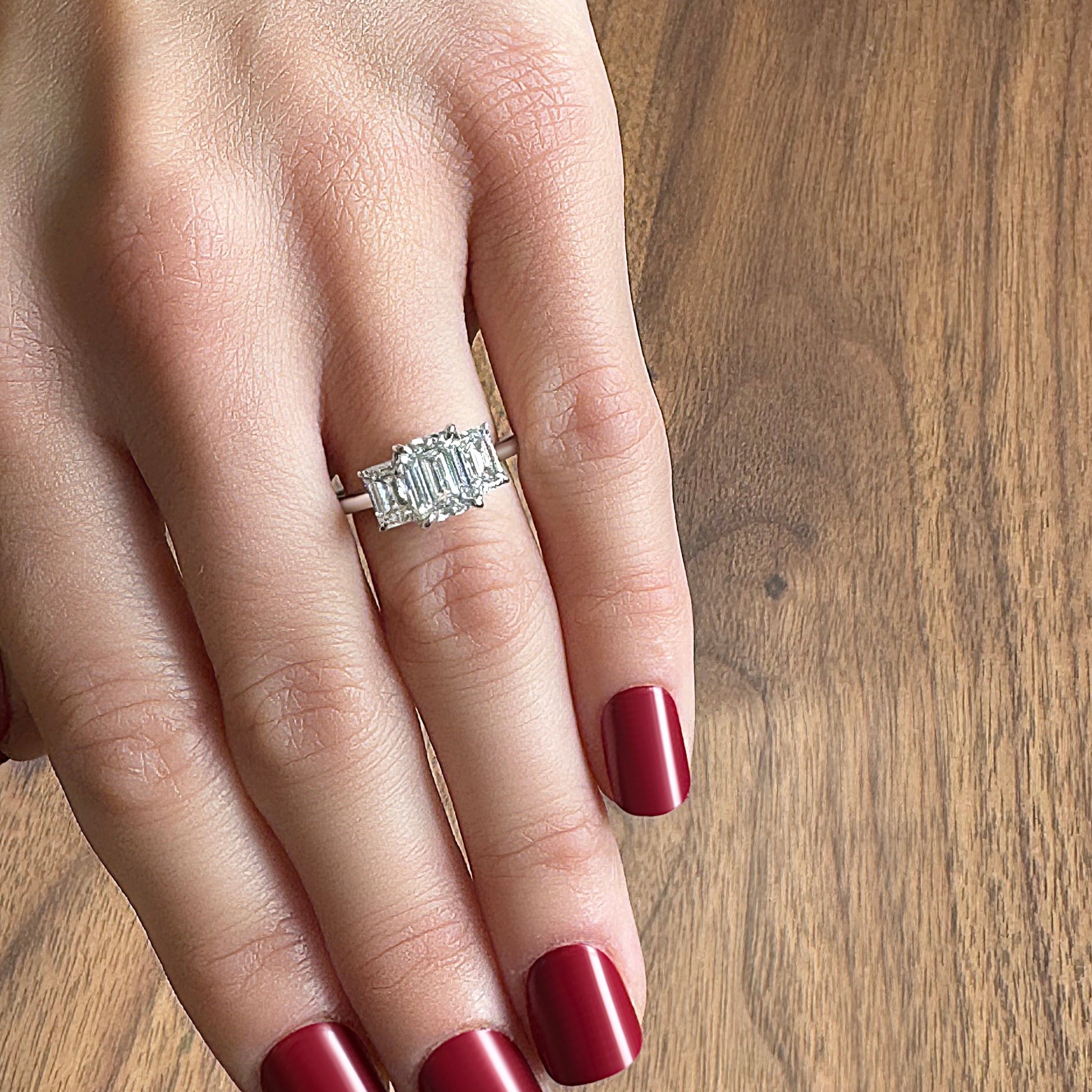 A hand with red manicured nails wearing a silver ring featuring three emerald-cut diamonds.