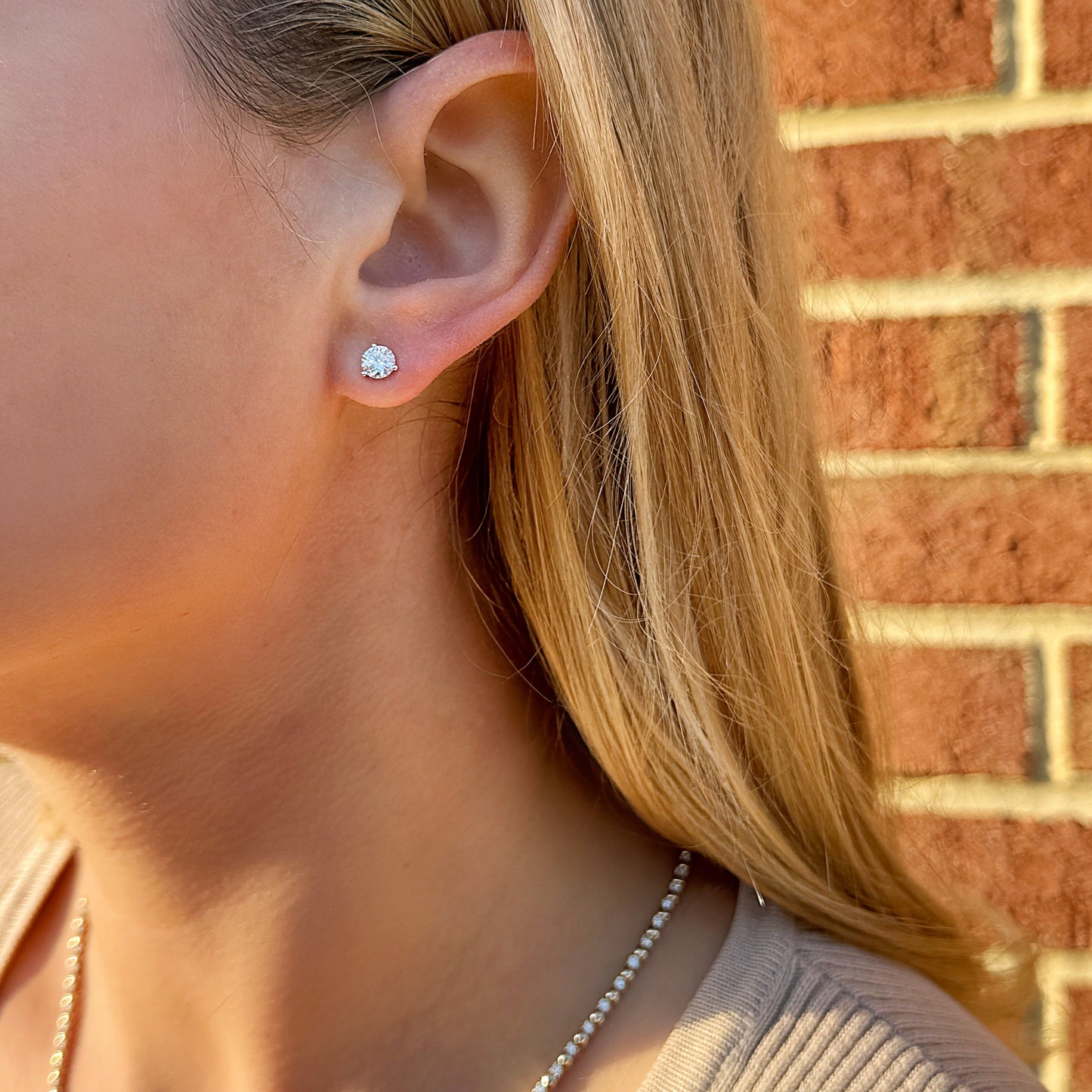 Close-up of a woman wearing a simple diamond stud earring against a brick background.