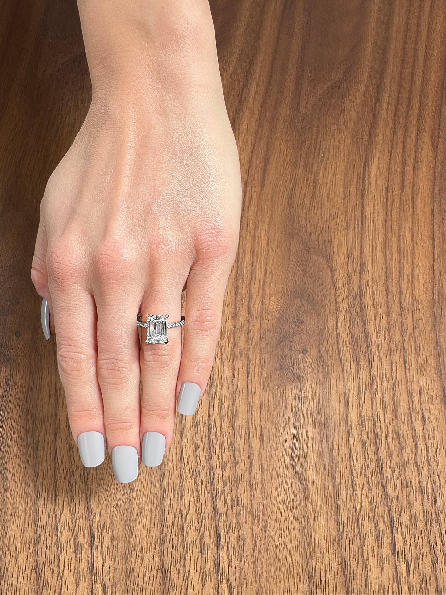 A hand with gray nail polish showcasing a diamond ring on a wooden surface.