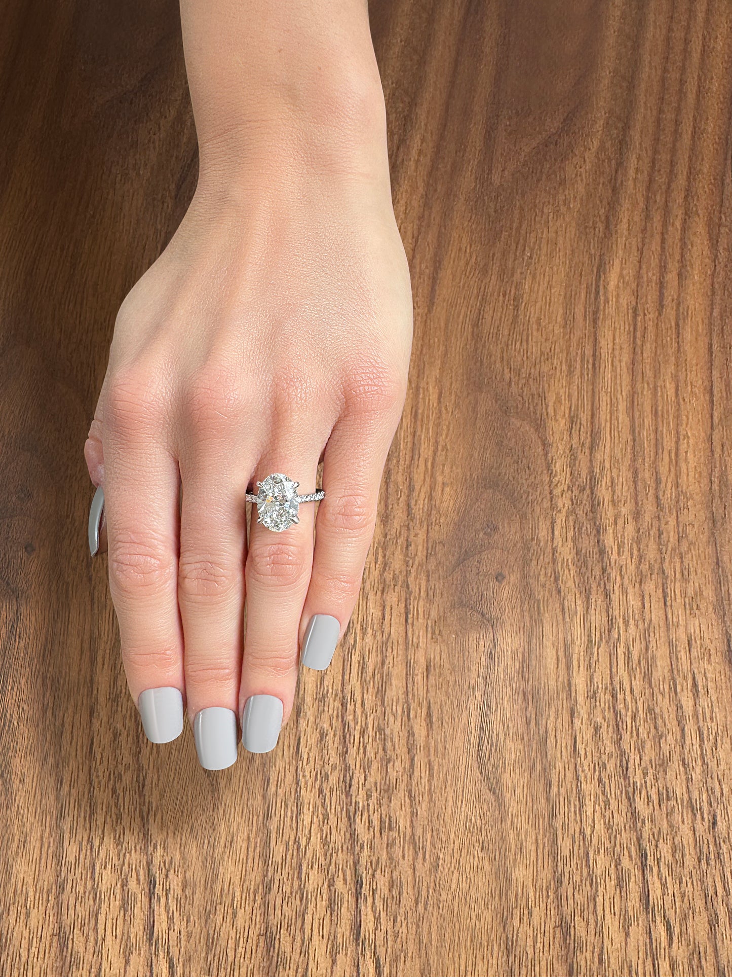 A hand with gray nails displays an elegant diamond engagement ring on a wooden surface.