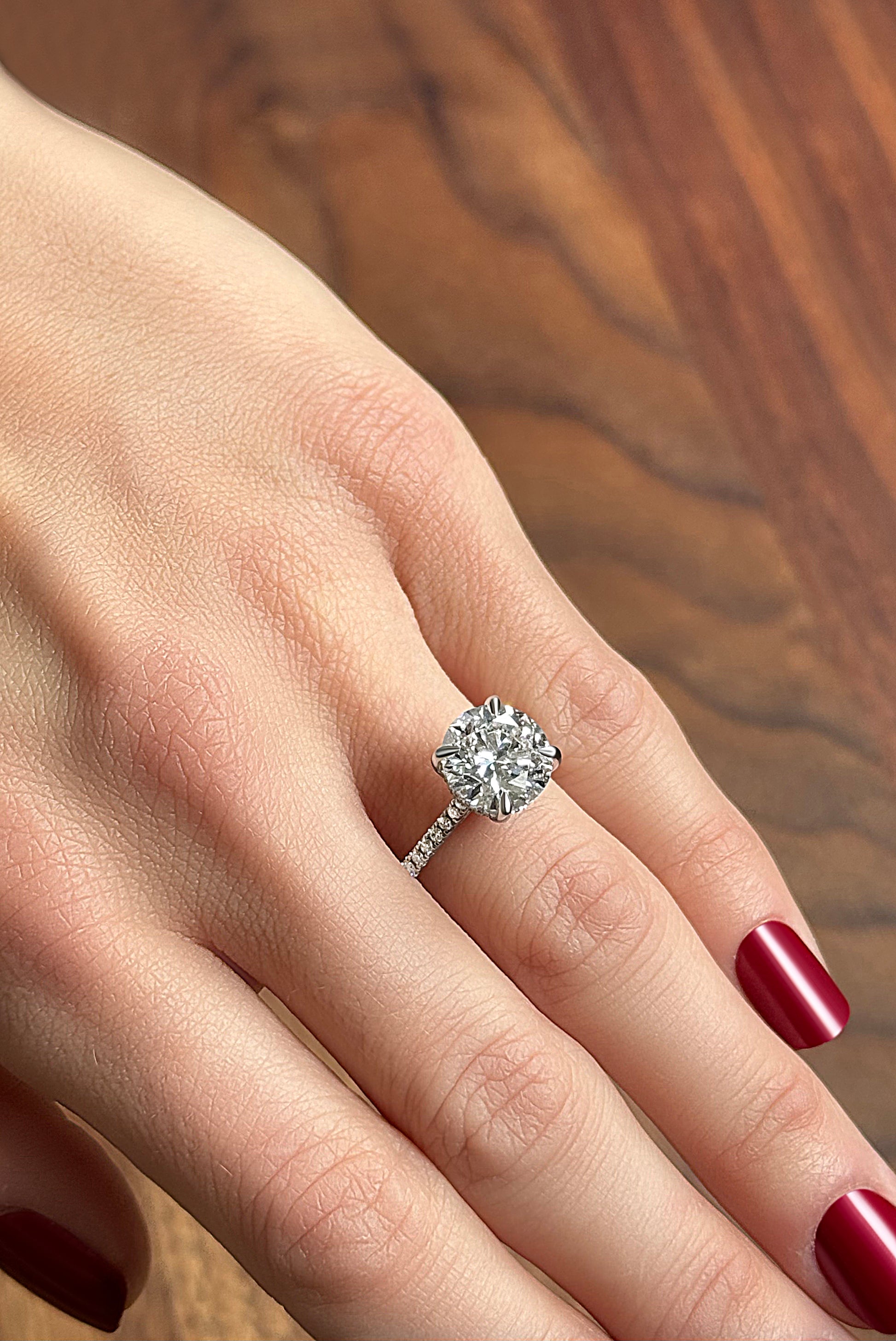 A hand with red nails wearing a sparkling diamond engagement ring on a wooden surface.