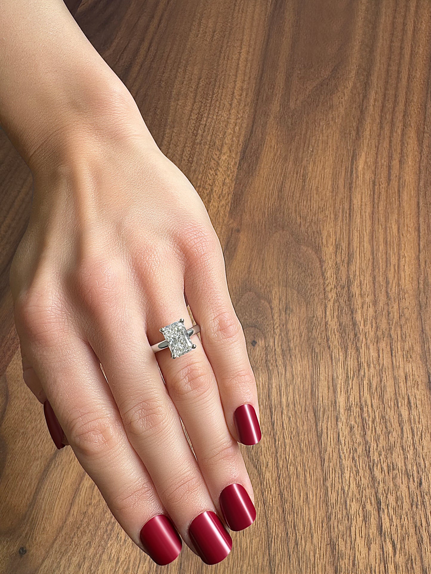A hand with deep red nails displaying a sparkling diamond ring on a wooden surface.