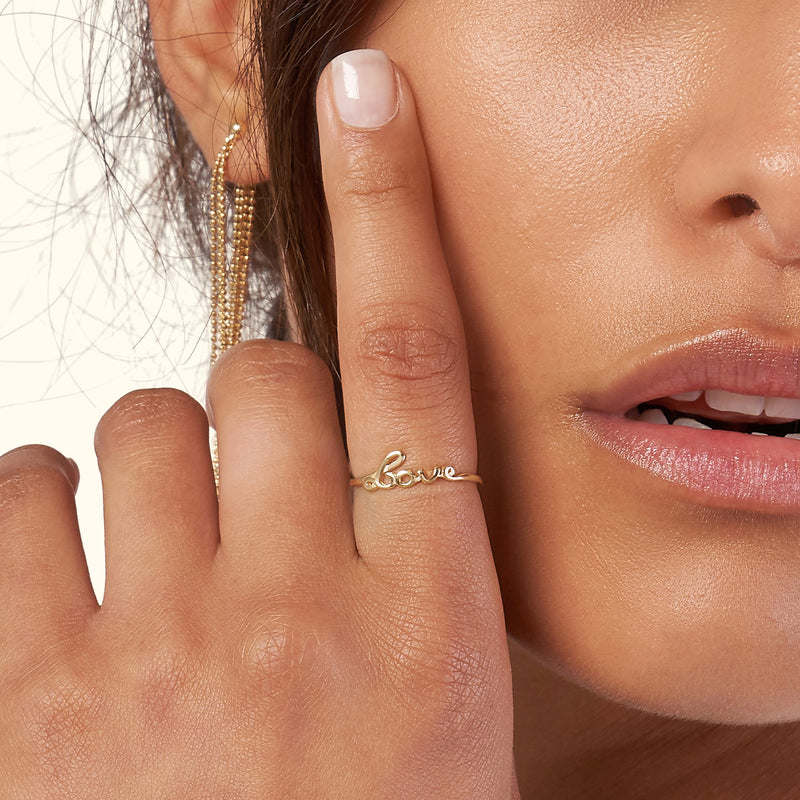 Gold ring shaped like the word "love" worn on a woman's finger beside her face.