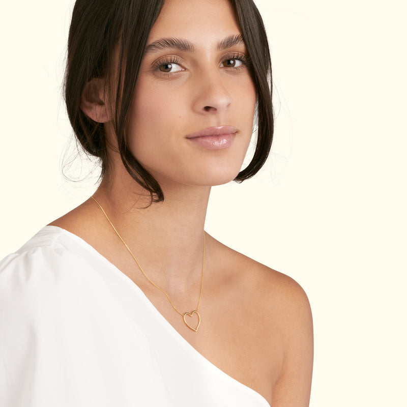 Model wearing a simple gold heart-shaped necklace, with a soft expression.
