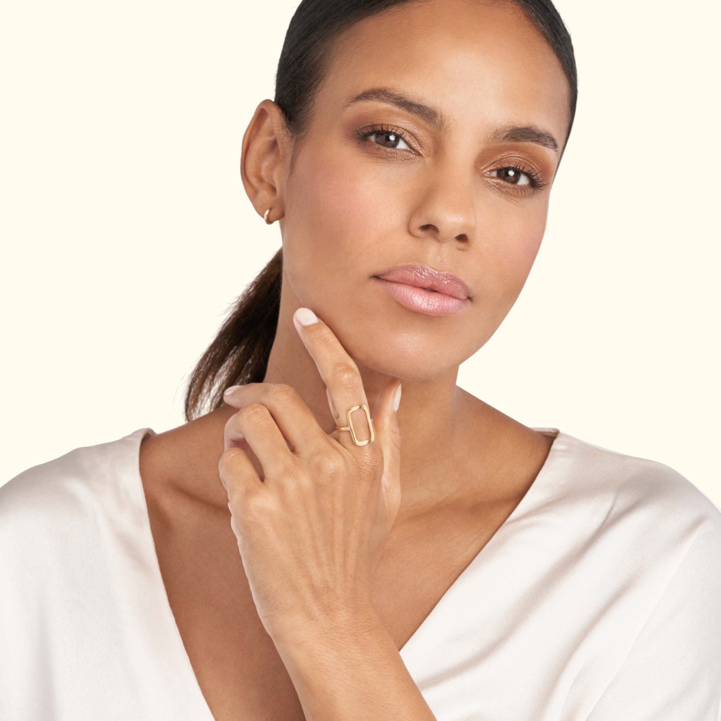 A woman in a white blouse gently touches a gold geometric ring on her finger.