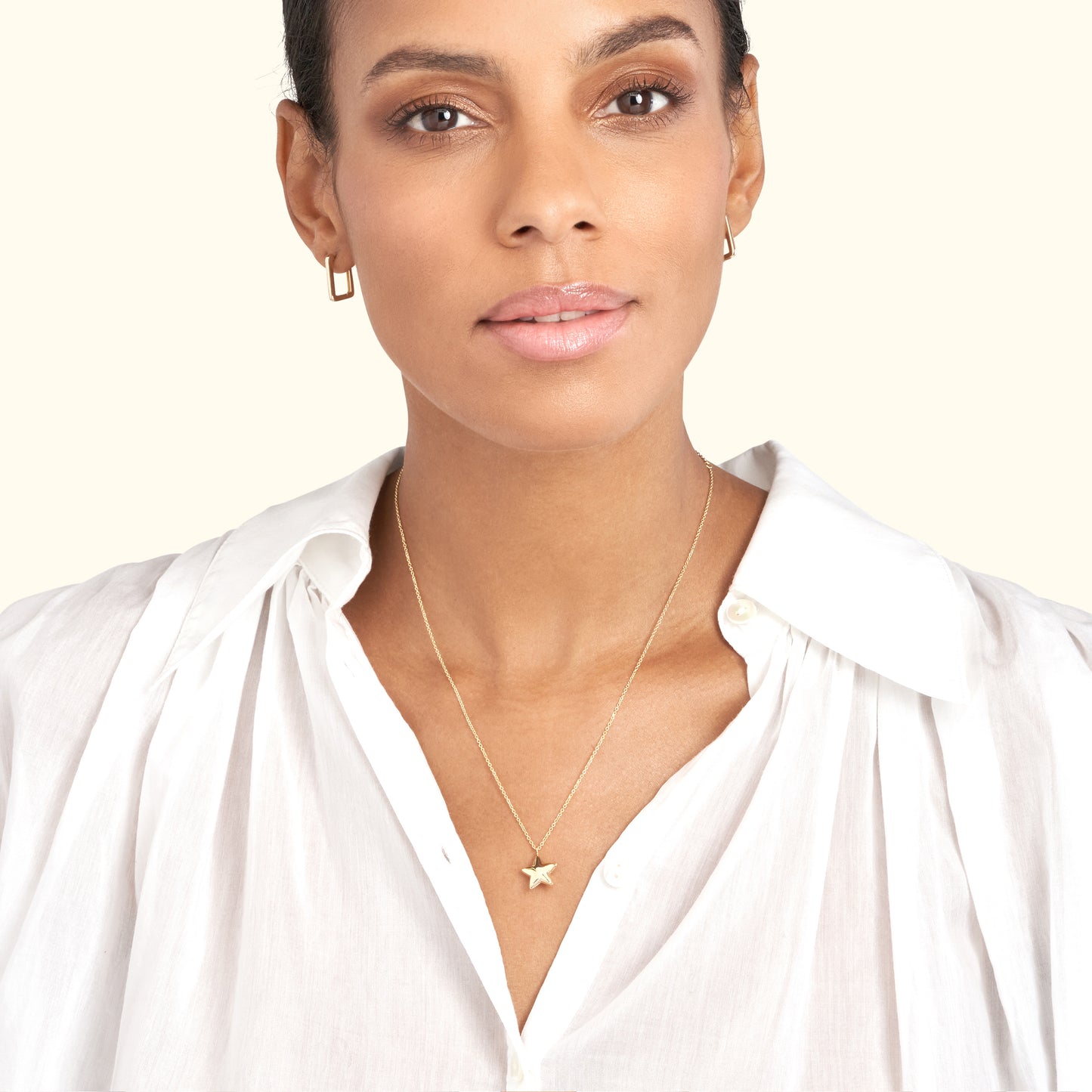 A woman wearing a gold star pendant necklace and small hoop earrings, smiling softly.