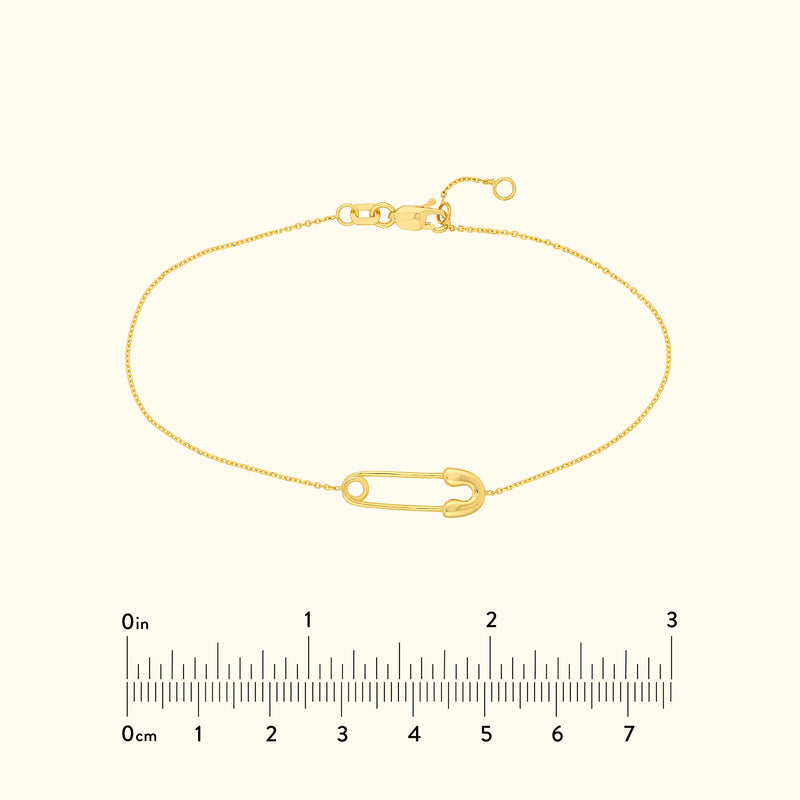 A delicate gold bracelet featuring a large safety pin charm, displayed with a ruler for size.
