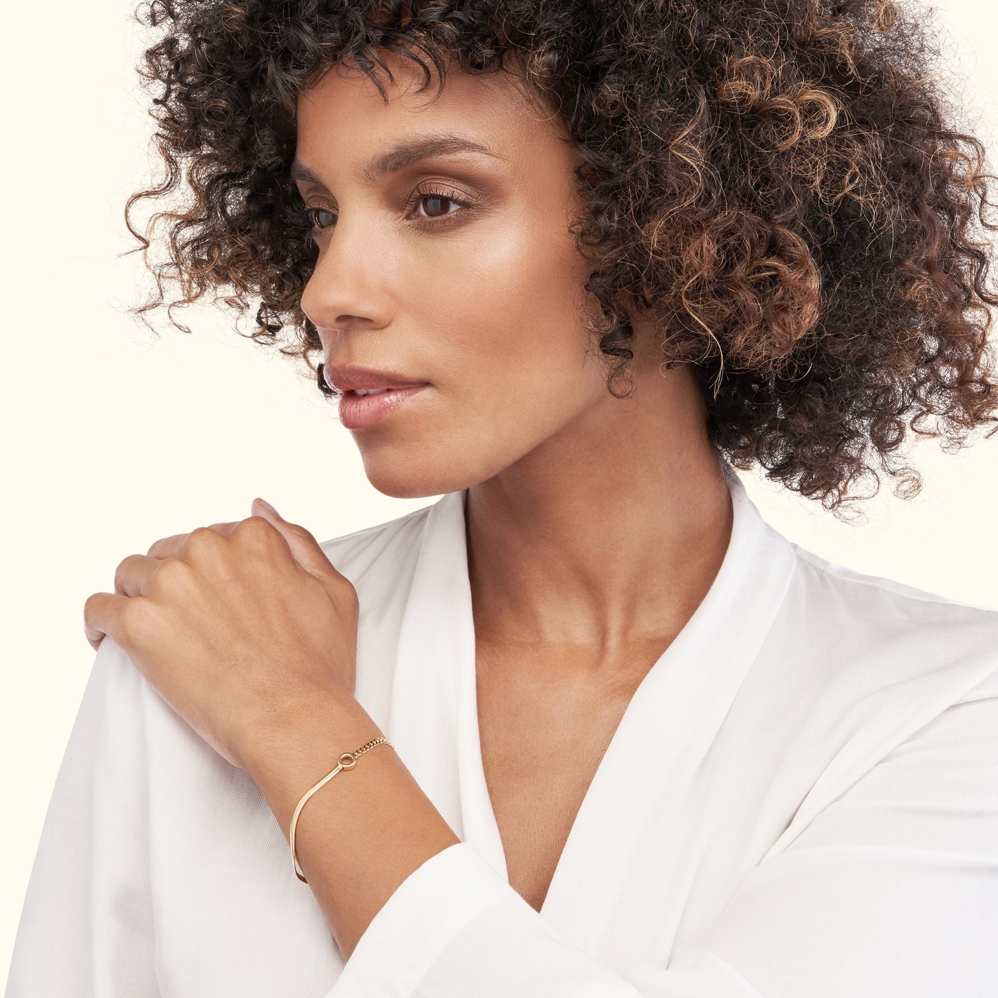 A woman with curly hair wearing a delicate gold bracelet, looking thoughtfully to the side.