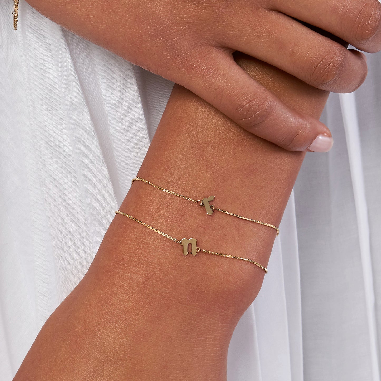 Two delicate gold bracelets with letter charms, worn on a wrist against a light fabric background.