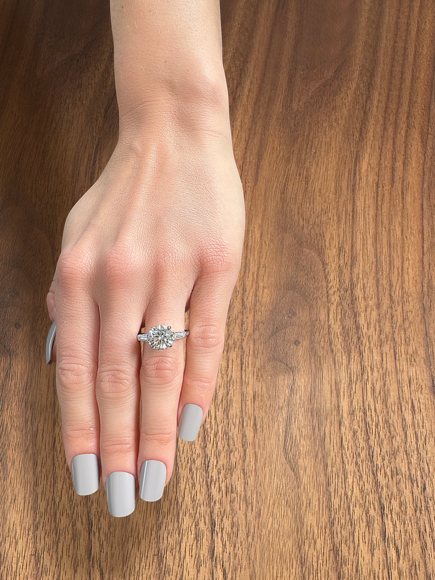 A hand with long, grey nails showcasing a sparkling diamond engagement ring on a wooden surface.