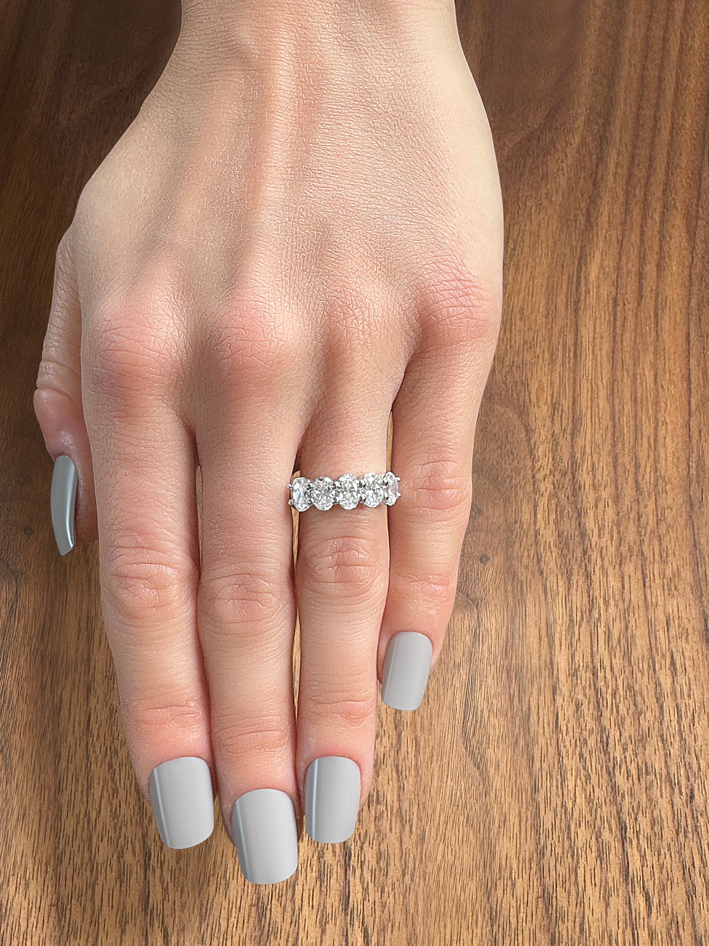 A hand with neatly manicured gray nails wearing a sparkling diamond band ring on a wooden surface.