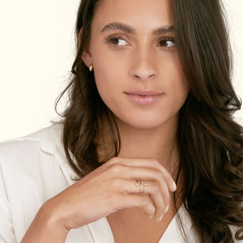 A woman with long brown hair wears a delicate gold ring and earrings, looking thoughtfully.