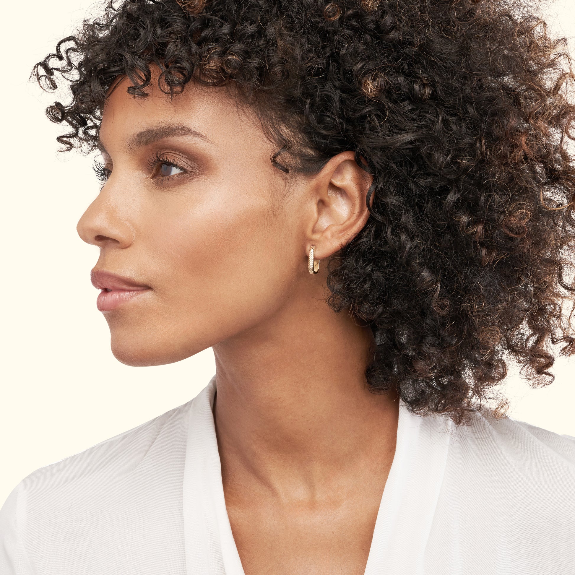 A profile view of a woman with curly hair wearing a small hoop earring and a white top.