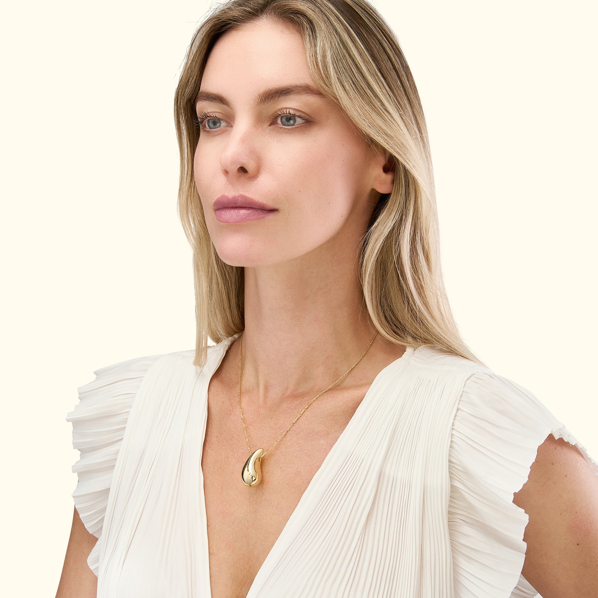 A woman wearing a gold pendant necklace, dressed in a white pleated top.