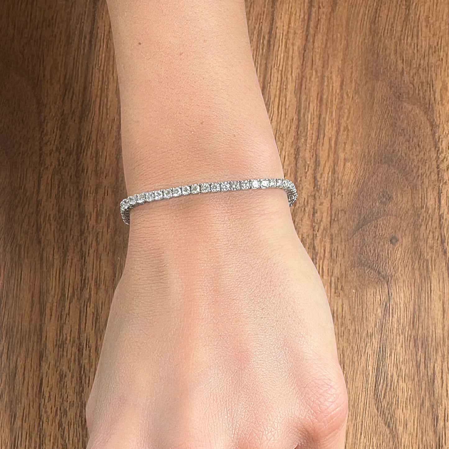 A hand wearing a delicate diamond tennis bracelet on a wooden surface.