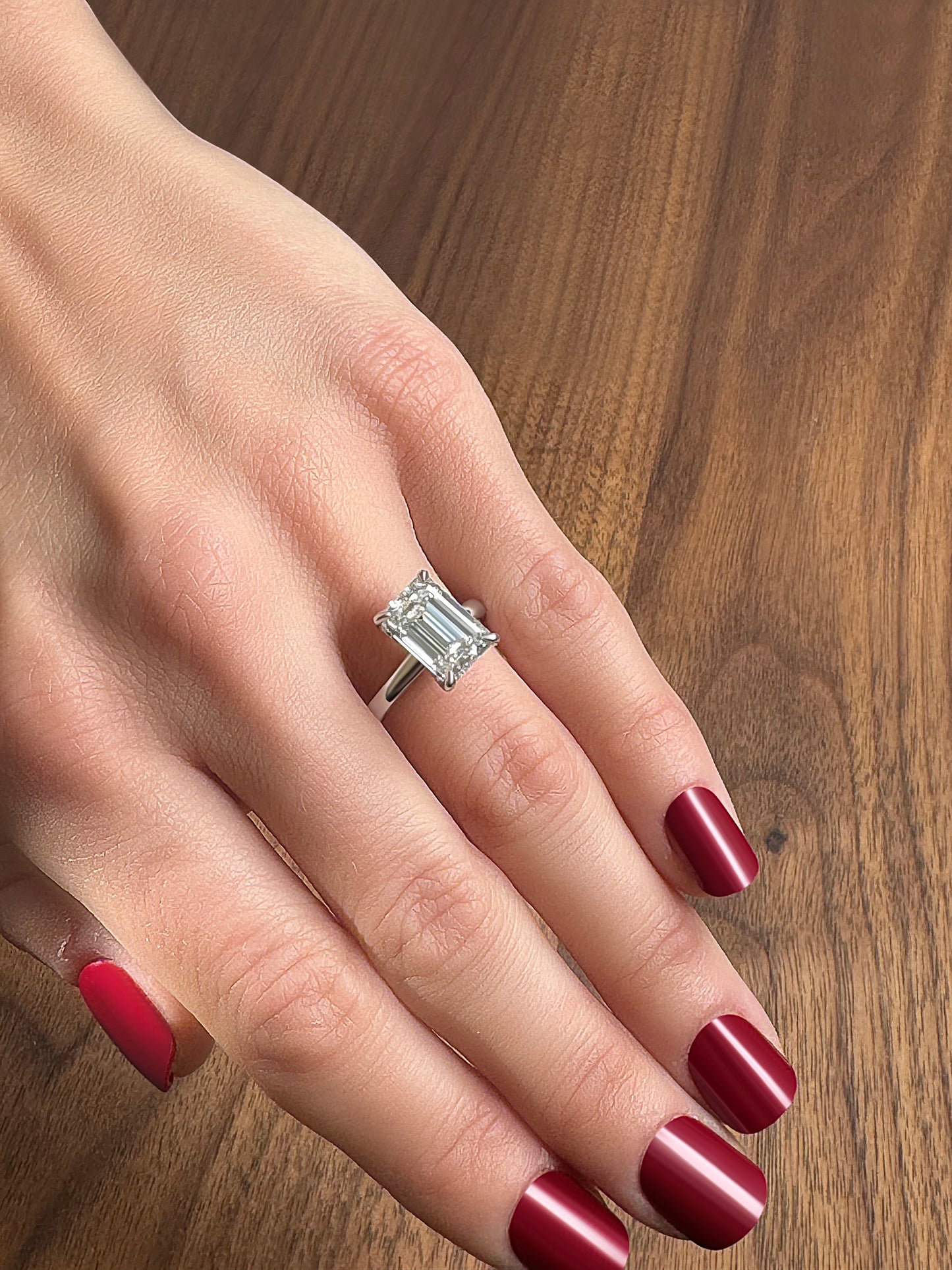 A hand with deep red nails showcasing a large emerald-cut diamond ring on a wooden surface.