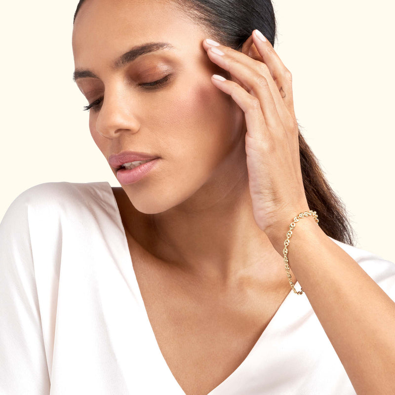 A woman wearing a delicate gold bracelet, gently touching her face, against a neutral background.