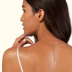 A woman with long hair wears a delicate gold necklace with a drop pendant, viewed from the side.