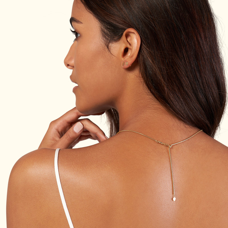 A woman with long hair wears a delicate gold necklace with a drop pendant, viewed from the side.