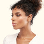 A woman with curly hair wears elegant gold hoop earrings, looking thoughtfully to the side.