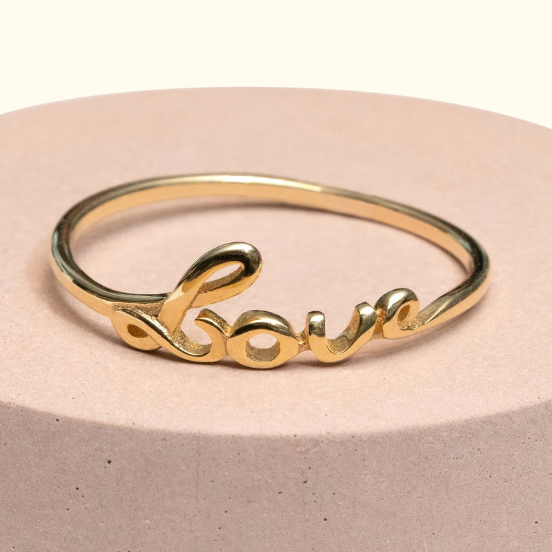 Gold ring shaped like the word "love" resting on a light-colored pedestal.