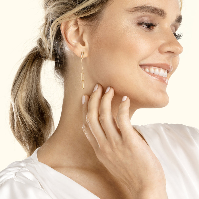 Smiling woman wearing gold paperclip earrings, gently touching her neck.