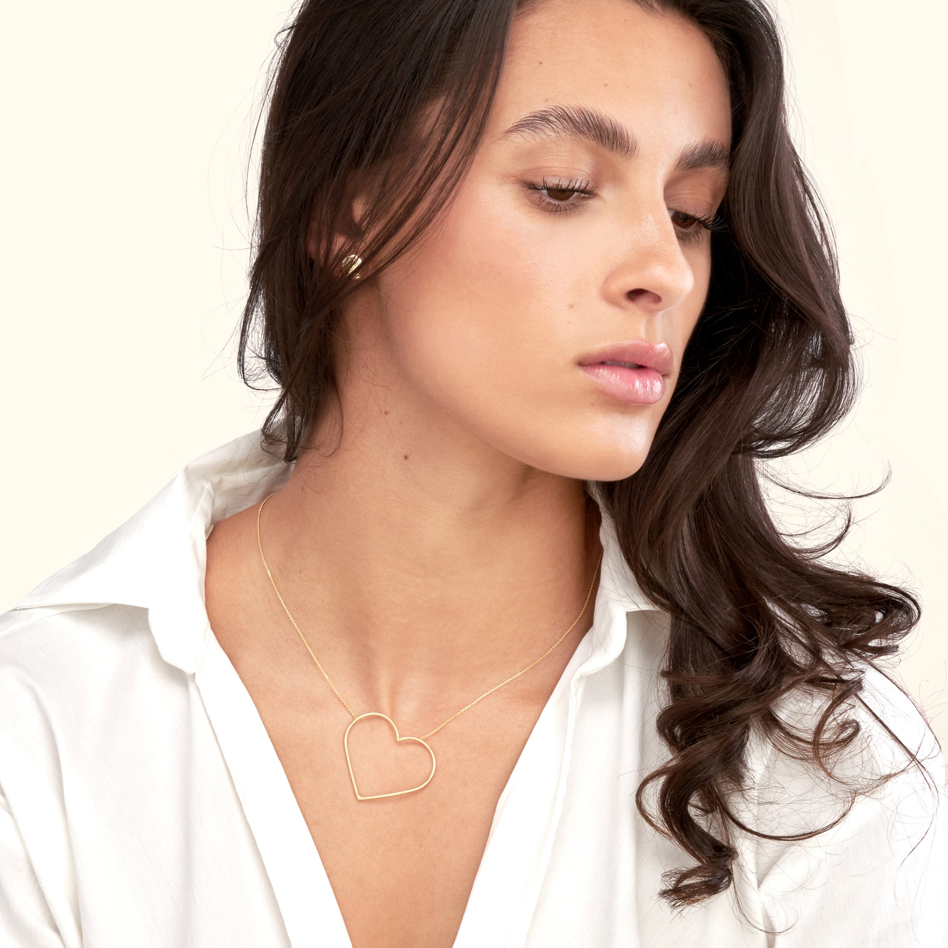 A woman with long hair wears a gold heart-shaped necklace, looking thoughtfully to the side.