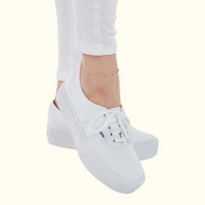 Delicate gold anklet worn with white canvas sneakers and ankle-length white jeans.