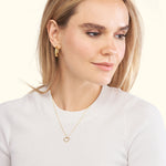 A woman wearing a gold heart-shaped necklace and hoop earrings, smiling gently.