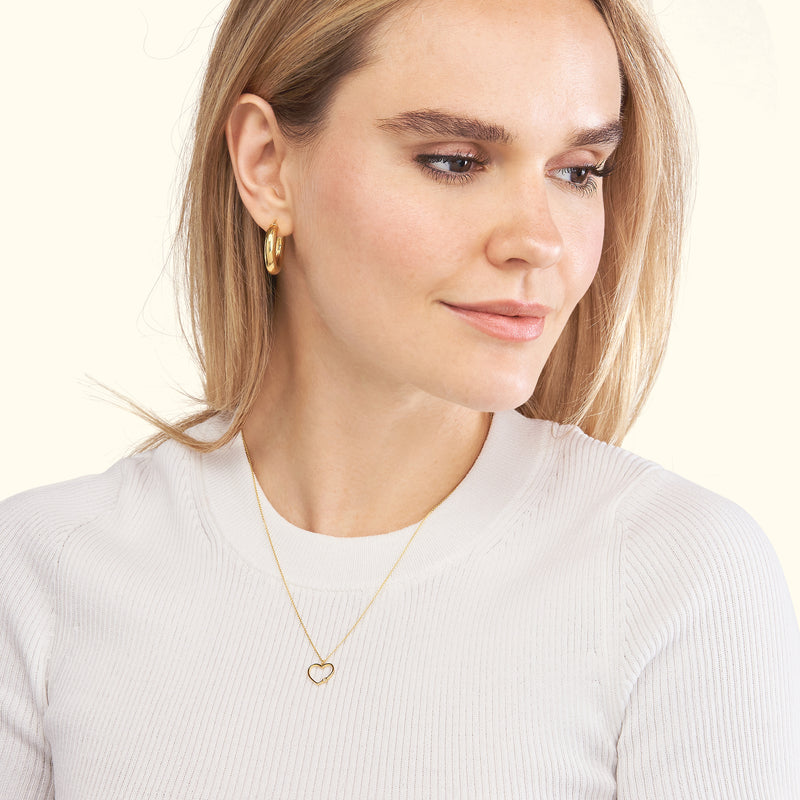A woman wearing a gold heart-shaped necklace and hoop earrings, smiling gently.