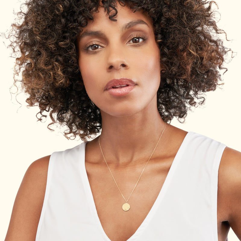 A woman with curly hair wearing a delicate gold necklace with a circular pendant.