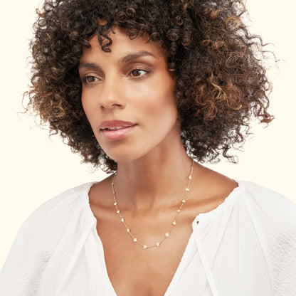 A woman with curly hair wears a delicate gold necklace featuring small, geometric charms.