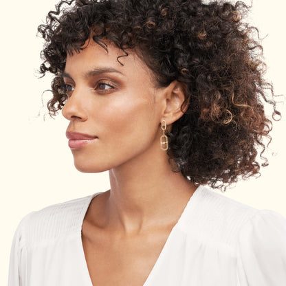 Gold geometric earrings worn by a woman with curly hair, dressed in a white blouse.