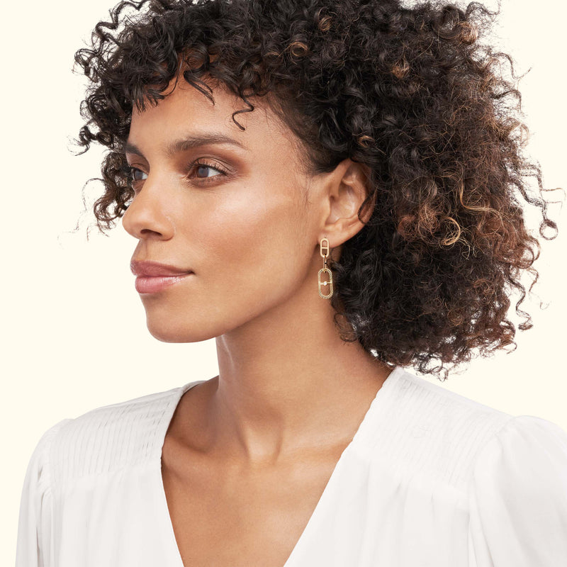 Gold geometric earrings worn by a woman with curly hair, dressed in a white blouse.