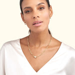 A woman models a gold chain necklace, wearing a silky white blouse against a light background.