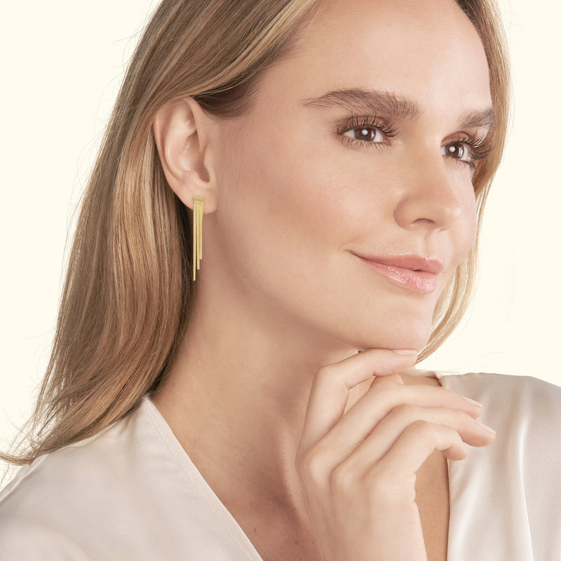 A woman with long hair wearing sleek gold earrings, smiling gently.