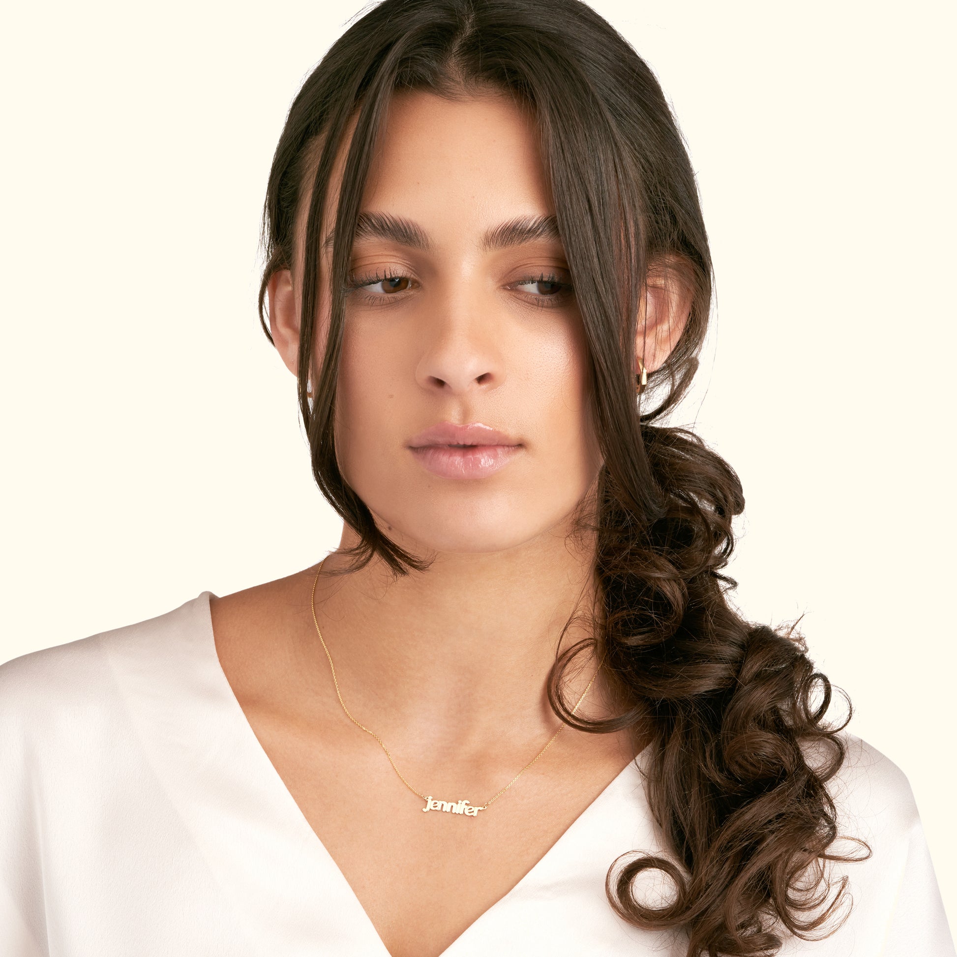 Young woman with long wavy hair wearing a gold name necklace, looking contemplative.