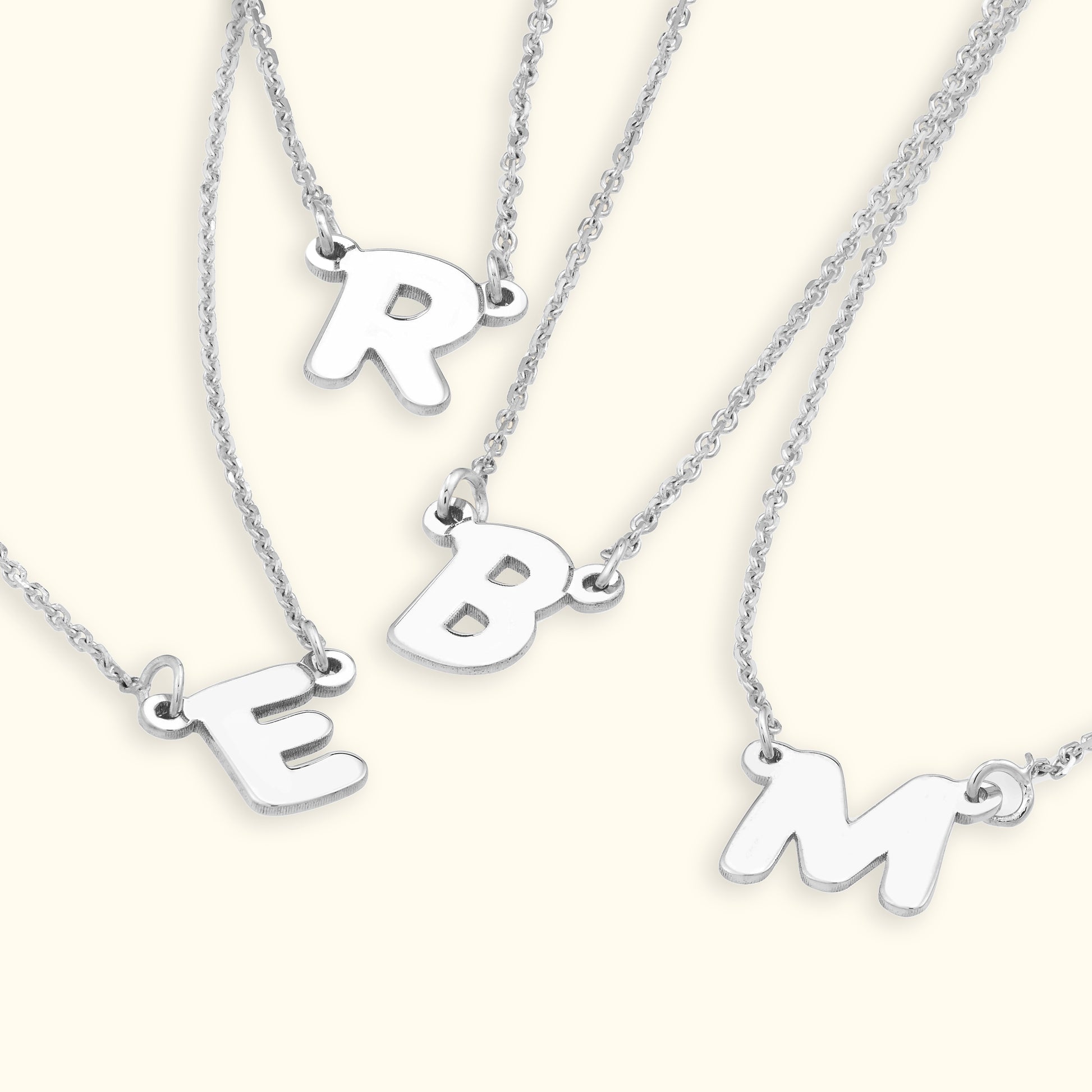 Four silver necklaces feature the letters R, E, B, and M hanging from delicate chains.