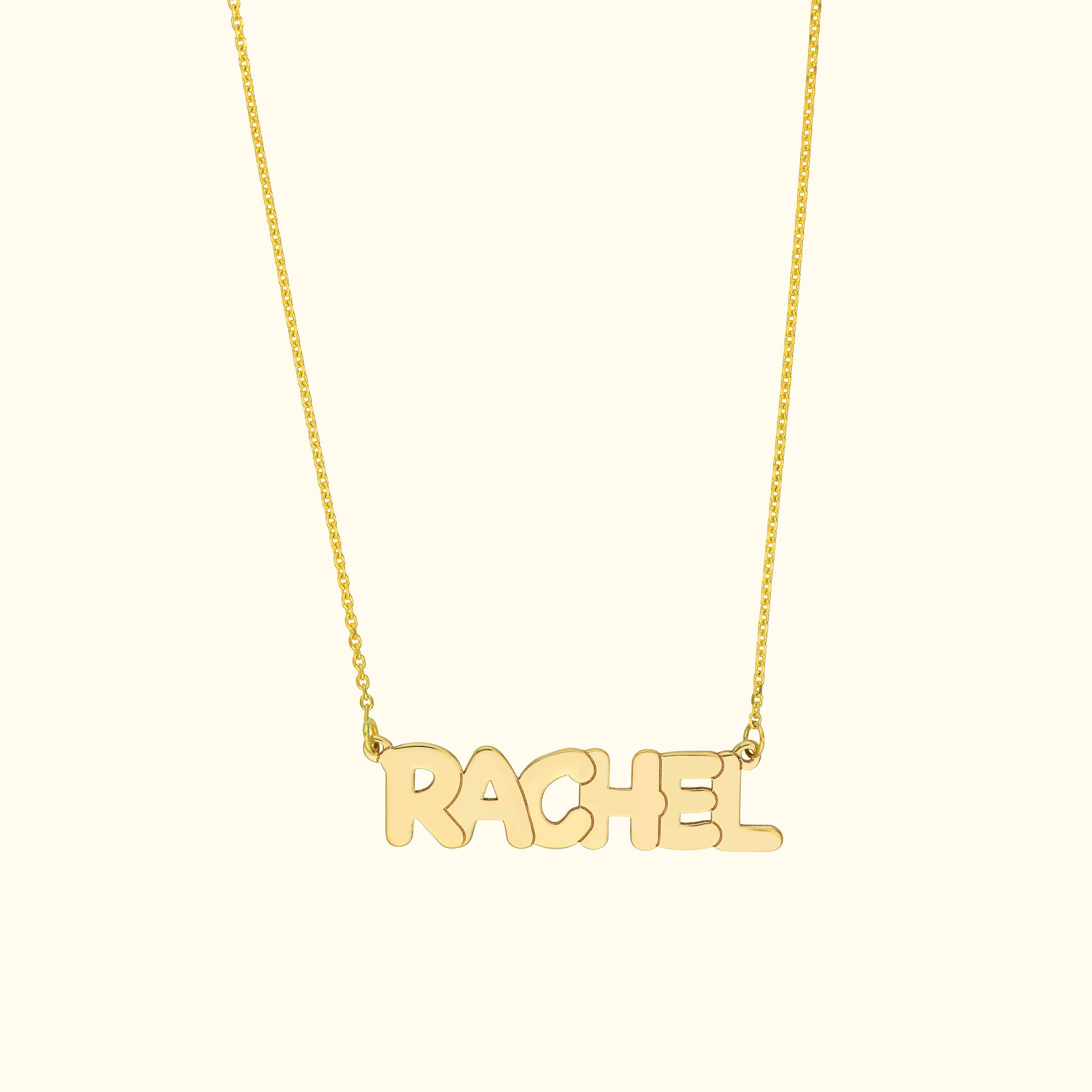 Gold name necklace spelling "RACHEL" on a delicate chain against a light background.