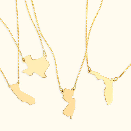 Four gold state-shaped pendants on delicate chains against a light background.