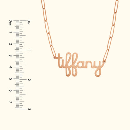 Rose gold necklace with cursive "tiffany" pendant, next to a ruler for size reference.