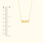 Gold necklace featuring the year "2024" as a pendant, displayed alongside a measuring ruler.