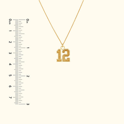 Gold necklace featuring a large, stylized number "12" pendant, with a ruler for size reference.