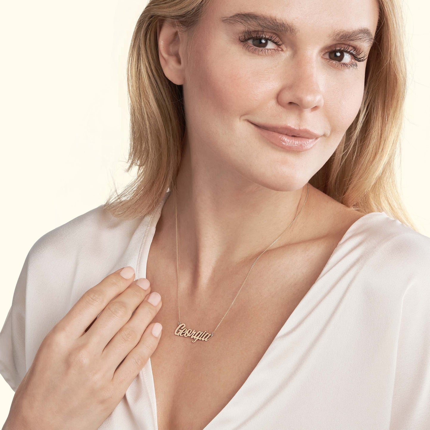 Woman in a white top holding a personalized gold name necklace, smiling gently.