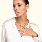 A woman wearing a gold chain necklace, lightly touching her neck, in a white blouse.