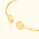 Gold cuff bracelet with minimalist round ends, set against a light beige background.
