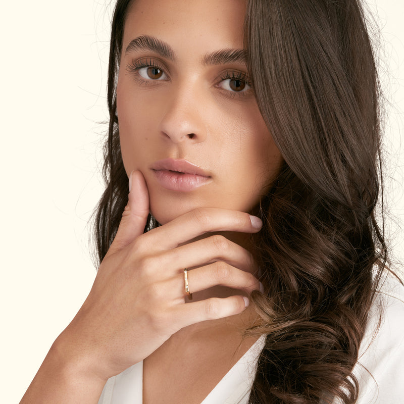 A woman with long hair gently touches her chin, showcasing a delicate gold ring.