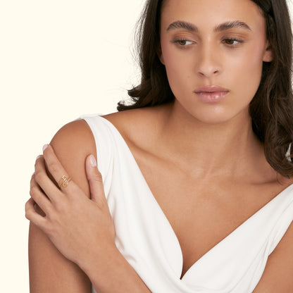 A woman in a white dress gently rests her hand on her shoulder, showcasing a delicate ring.