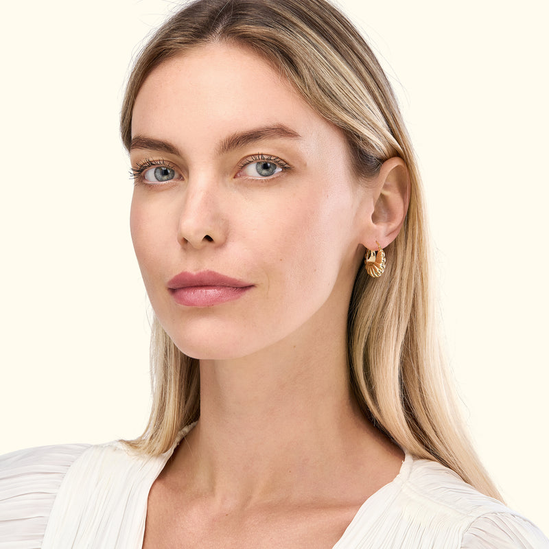 A woman with long blonde hair is wearing gold hoop earrings and a white blouse.